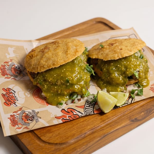 Two gorditas with green salsa and lime wedges on branded paper, served on a wooden board.