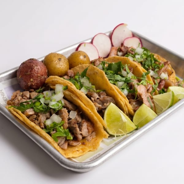 Four street tacos with meat, cilantro, and onions, served with limes, radishes, and potatoes on a tray.
