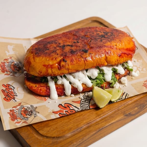 A large, red-orange Mexican torta sandwich with white sauce, lettuce, and cheese, served with lime wedges.