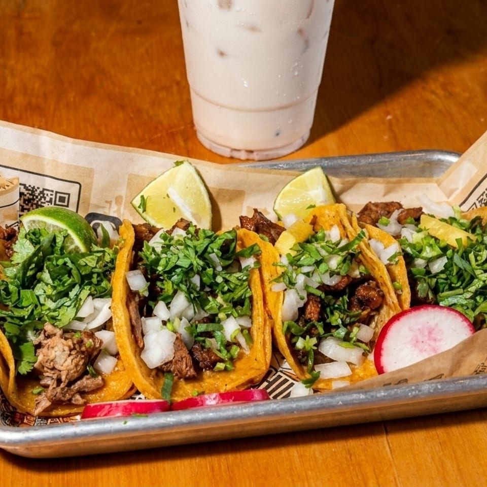 Five street tacos with meat, onions, cilantro, lime, and radish on a tray with a drink.