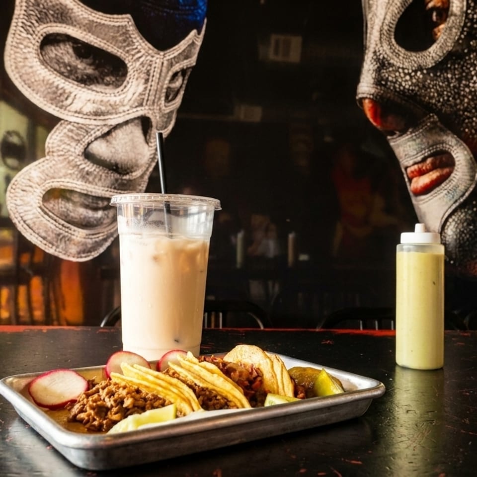 Tacos, a drink, and sauces on a table with lucha libre masks painted on the wall.