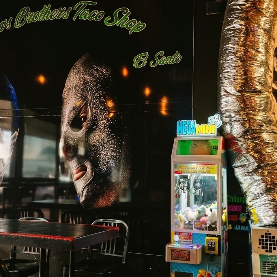 Interior of Chilangos Brothers Taco Shop with wrestling mask murals, a claw machine, and black tables.