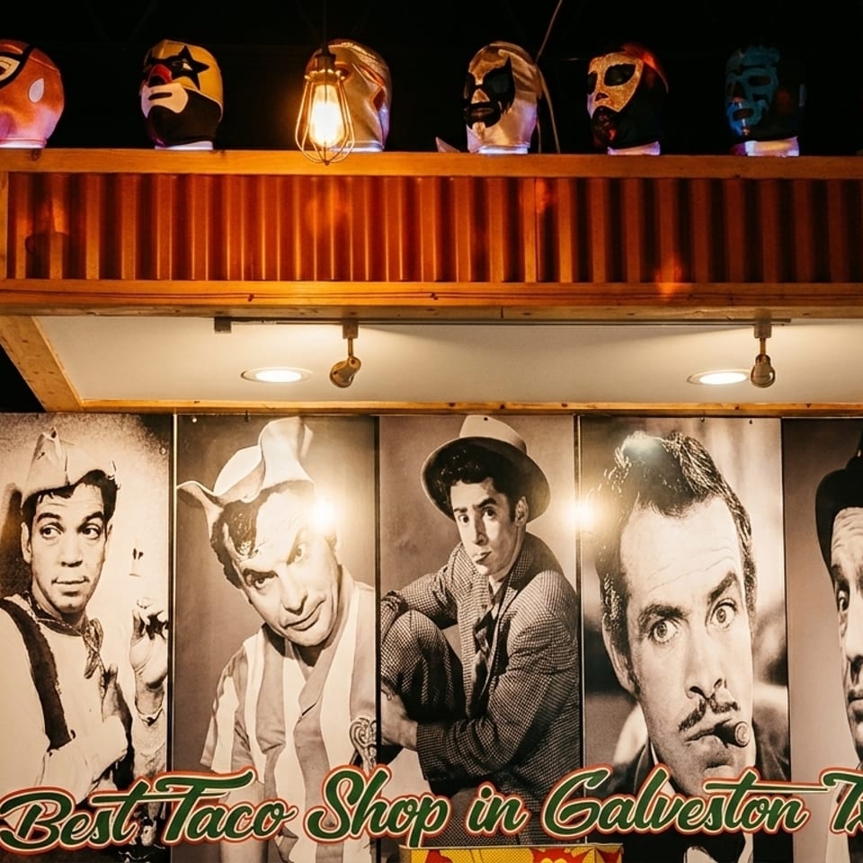 Lucha libre masks on a shelf above black and white portraits of men, with "Best Taco Shop in Galveston Tx" text.